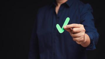 Close-up of hand holding a green check mark symbol while standing on a black background photo