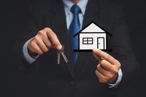 Business and real estate concept. A businessman in a suit holding keys and a mini paper house while standing in the office photo