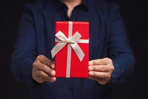 Close-up of hands holding a red gift box with a white ribbon while standing on a black background photo