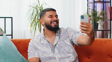 Happy Indian man taking selfie on smartphone communicating call home online with subscribers video