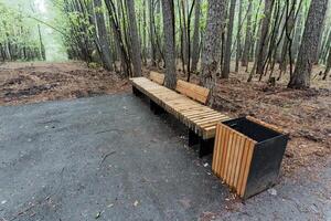 A strong wooden bench next to a trash can in a tranquil forest is the perfect spot to sit, relax, and admire the breathtaking beauty of nature, making it an ideal retreat photo