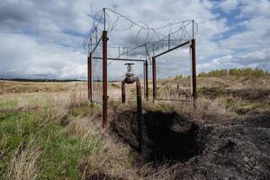 Abandoned Infrastructure in a Rural Landscape A disused rustic pipeline entwined with a barbed wire structure stands starkly against the backdrop of an empty field framed by the vast sky above photo
