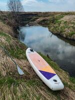A paddle board along with a paddle is positioned next to a gently flowing river, surrounded by the natural beauty of the landscape, ready for an exciting adventure on the water photo