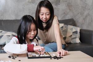 Single mother, and child concentrate playing Go, checkers board game on table in home living room together with fun and cheerful, happy weekend relaxing hobby of Asian family. photo