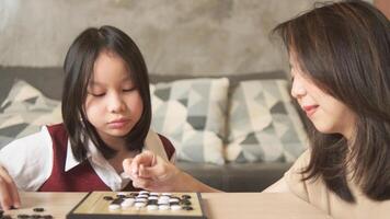 Asian family, child, and mother cheerful playing Go, checkers board game on table in home living room together, happy weekend relaxing hobby of kid and parent, concentrate and compete. photo