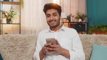 Indian man on sofa smiling giving thumbs up showing like gesture looking at smartphone feeling happy video