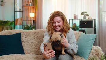 Young woman waving at call on smartphone with pug dog on home sofa, joyful friendly greeting video