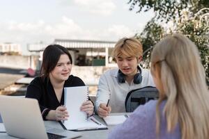 Diverse Group of Young Students Collaborating and Studying Together Outdoors with Laptops and Notebooks in a Modern Educational Setting photo