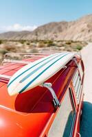 Surfboard resting on the roof of a vibrant red vintage vehicle, surrounded by arid landscapes and mountains, evoking a sense of adventure and summer vibes photo