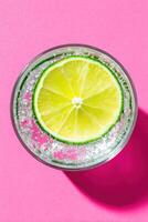 Refreshing beverage in a clear glass with a slice of lime floating on top, surrounded by bubbles, set against a vibrant pink background, evoking summer vibes and thirst-quenching moments photo
