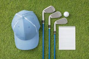 Golfing accessories including a blue cap, three golf clubs, a golf ball, and a notepad arranged neatly on vibrant green grass, representing the sport and preparation for a game photo