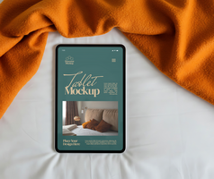 A tablet displaying a mockup design rests on a bed under a warm orange blanket psd