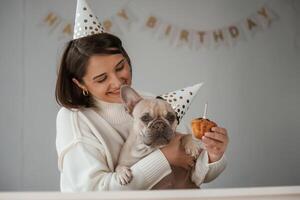 Birthday of the pet. In festive ,hats, and with cake. Young woman is with her pug dog at home photo
