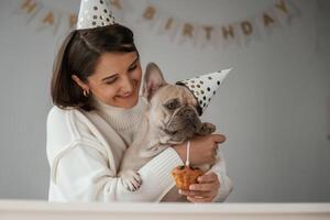 Birthday of the pet. In festive hats, and with cake. Young woman is with her pug dog at home photo