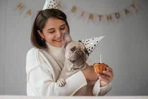 Birthday of the pet. In festive, hats and with cake. Young woman is with her pug dog at home photo