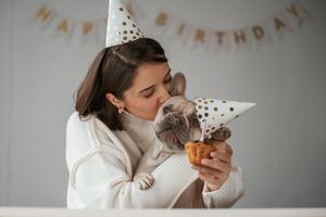 Happy owner is celebrating, birthday of pet. In hat and with cake. Young woman is with her pug dog at home photo