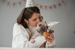 Happy, owner, is celebrating birthday ,of pet. In hat and with cake. Young woman is with her pug dog at home photo