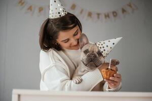 Happy, owner, is celebrating ,,birthday of pet. In hat and with cake. Young woman is with her pug dog at home photo