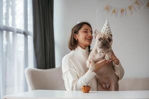 By, the table, celebrating happy birthday of pet. Young woman is with her pug dog at home photo
