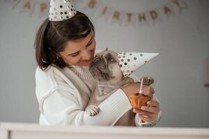 Happy, owner is celebrating birthday of pet. In hat and with cake. Young woman is with her pug dog at home photo