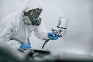 In protective mask,, and wear. Close up view of man that is painting a car by using spray guy photo