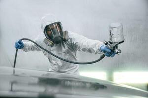 In, protective mask and wear. Close up view of man that is painting a car by using spray guy photo