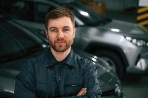 With, arms crossed. Repairman in uniform is standing in the garage with cars photo