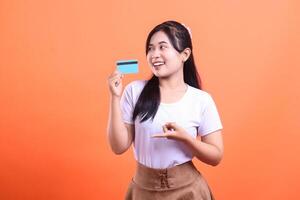 Young smiling woman holding credit card and pointing on it with a finger. She is delighted with her shopping plan, isolated on orange background photo