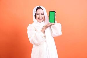 A smiling Muslim woman with a hijab is showcasing a smartphone with a green screen. Ideal for Ramadan-themed advertisements. isolated on a orange background. photo