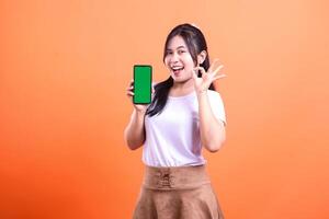 Smiling woman showing a smartphone with a green screen, okay hand gesture on orange background photo