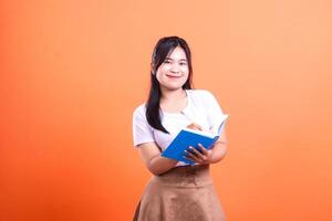 A young woman is writing in a blue notebook with a pen while standing on a orange background photo