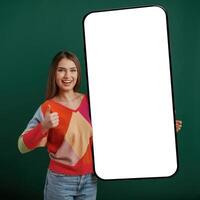 Large, smartphone. White display for text or design. Young woman is standing against green background in the studio photo