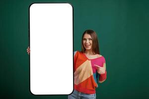 Large smartphone. White ,display ,for text or design. Young woman is standing against green background in the studio photo