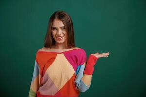 Showing, by ,hand, to the side, place for text. Young woman is standing against green background in the studio photo