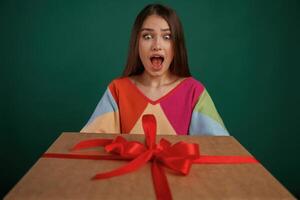 Surprised, holding ,box ,gift box. Young woman is standing against green background in the studio photo