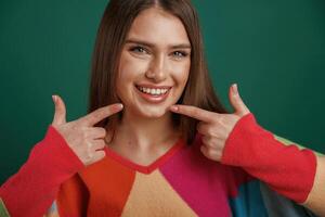 Pointing to the teeth by fingers, smiling. Young woman is standing against green background in the studio photo