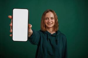 White display of smartphone,, place for text. Young woman is standing against green background in the studio photo