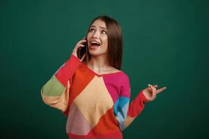 Talking, by phone, having conversation. Young woman is standing against green background in the studio photo