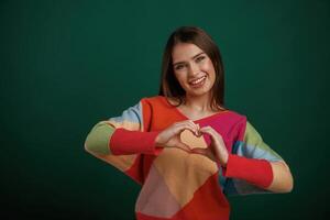 Heart shaped gesture by hands, sending love. Young woman is standing against green background in the studio photo