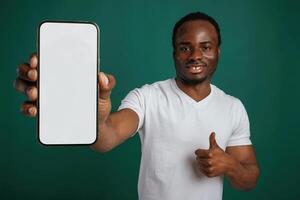 Smartphone ,with white empty space for text. African American man is standing against green background in the studio photo