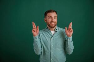 Fingers crossed, hoping for better. Man is standing against background in the studio photo