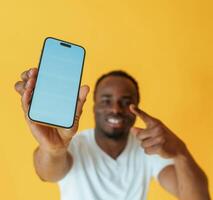Blue colored smartphone for copy space in hand. Black man is in the studio against yellow background photo