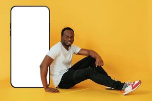 Sitting near to the ,,large ,smartphone with copy space. Black man is in the studio against yellow background photo