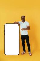 Leaning ,on the large ,smartphone for copy space. Black man is in the studio against yellow background photo