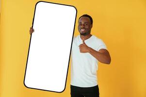 With very big smartphone ,for ,,copy space.. Black man is in the studio against yellow background photo