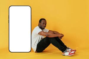 Sitting near ,to the large smartphone ,with copy space. Black man is in the studio against yellow background photo