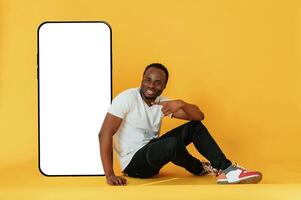 Sitting near, to,, the large smartphone with copy space. Black man is in the studio against yellow background photo