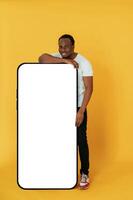Leaning on the large smartphone ,for, copy space. Black man is in the studio against yellow background photo