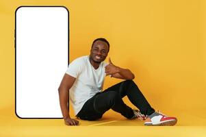 Sitting near to ,the large smartphone ,with copy space. Black man is in the studio against yellow background photo