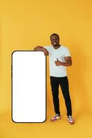 Leaning on the large smartphone ,for copy space. Black man is in the studio against yellow background photo